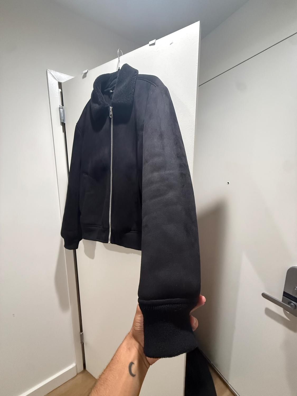Here’s the rephrased sentence: A hand holds a dark outer garment, fitted with a zipper closure and a velvet-touch collar, which is suspended above a white door.