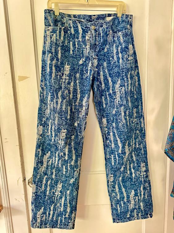 Blue denim jeans with a distressed design and abstract pattern hanging on a hanger over a white door.