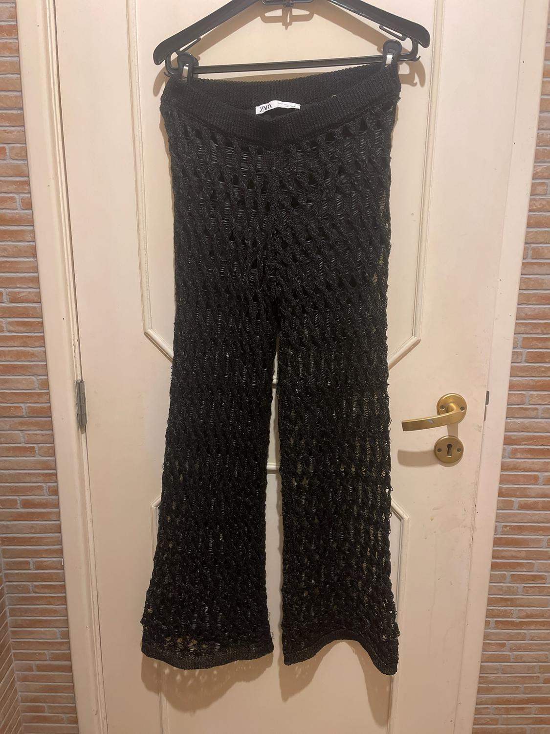 Black open-knit trousers hanging on a hanger over a white door. ZARA label visible.