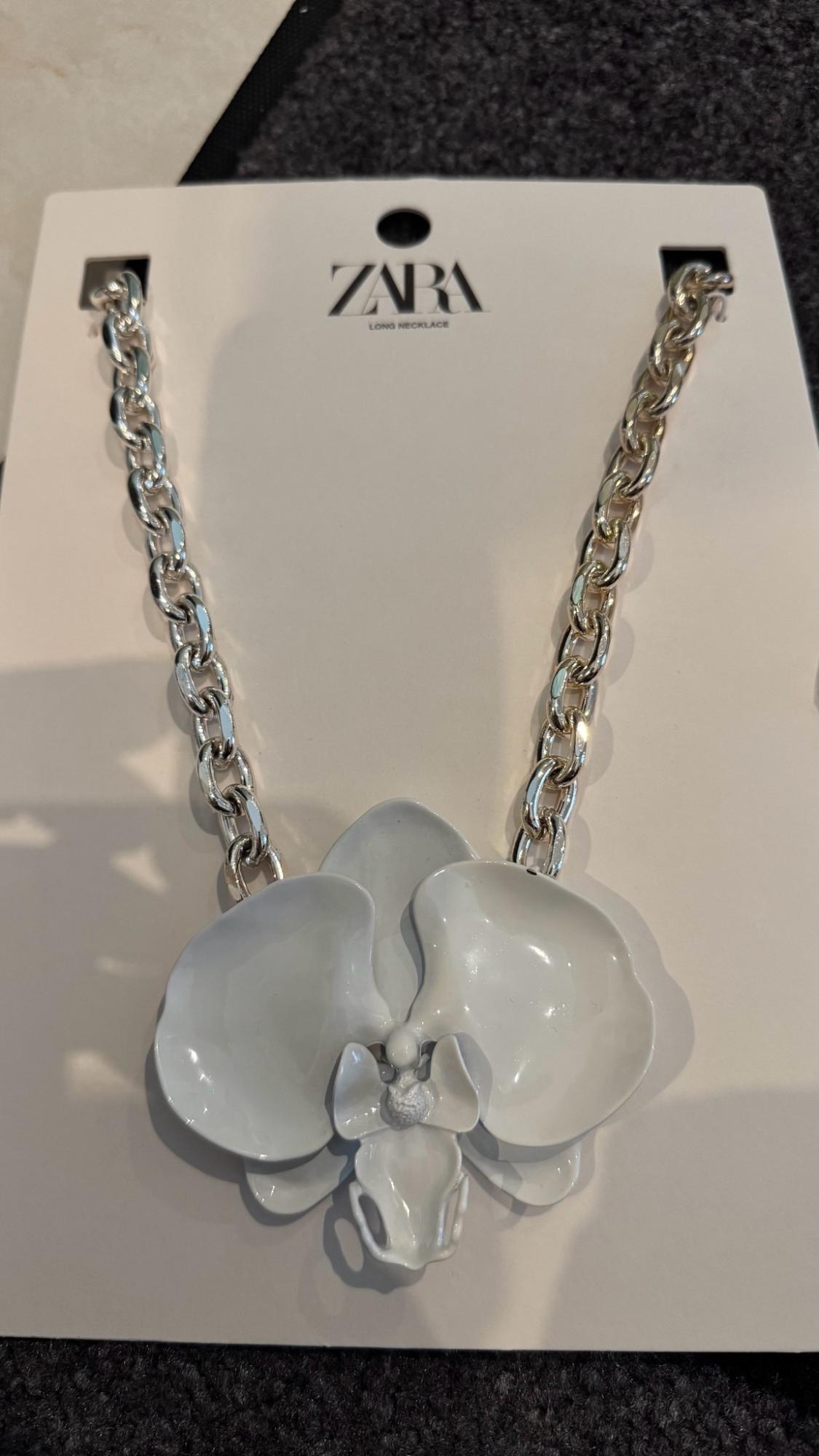 Silver-coloured chain necklace with a white flower pendant on a tag reading ZARA LONG NECKLACE.