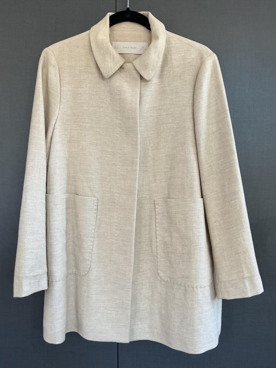 Light beige coat hanging on a black hanger against a dark background. It features a collar, a concealed front closure, and two pockets. ZARA BASIC label.