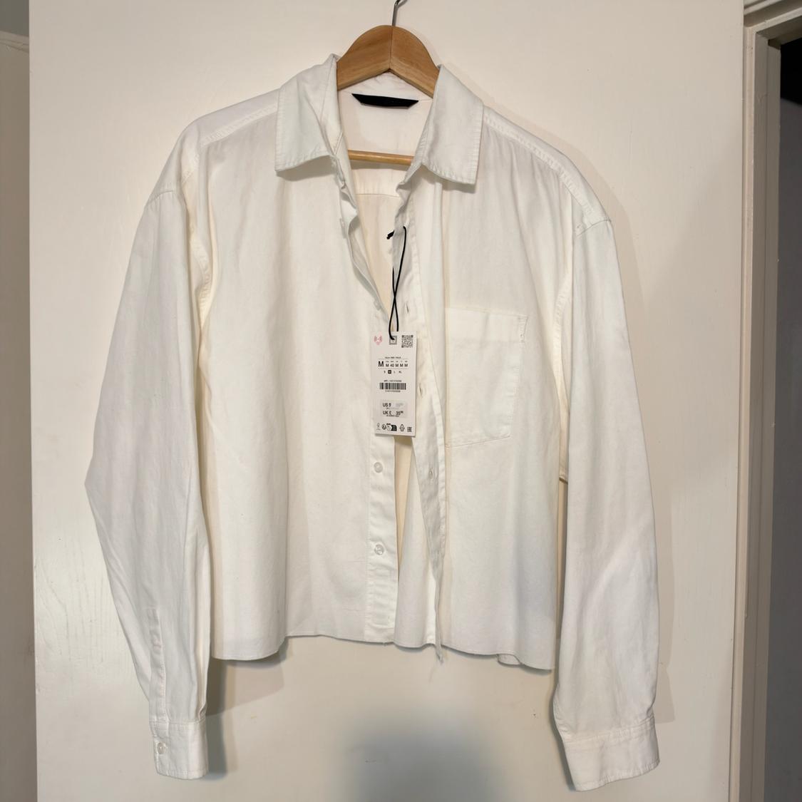 Relaxed-fit white shirt, long sleeves with a front pocket, hanging on a wooden hanger on a white door.