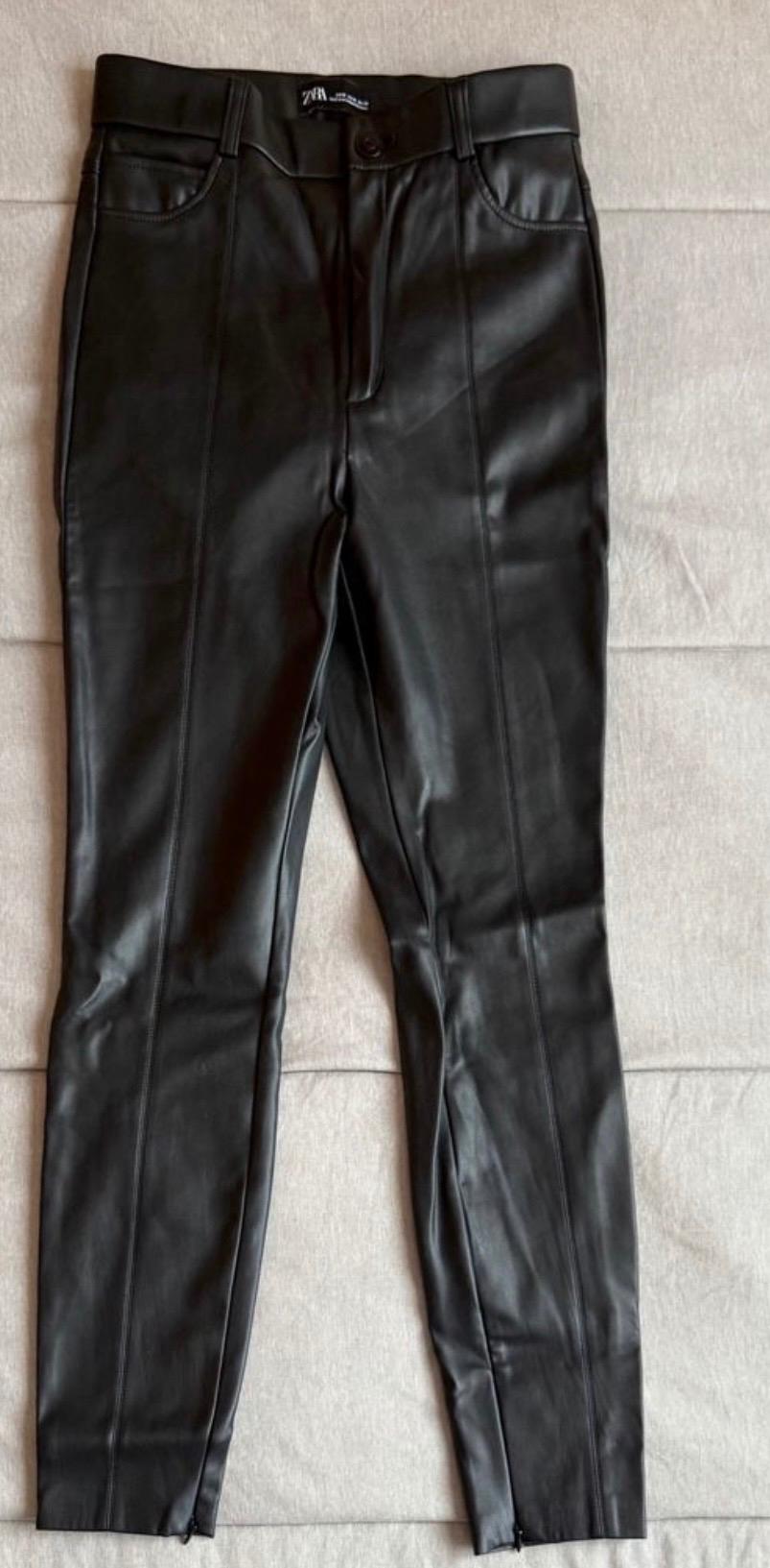 Black trousers made from synthetic material with visible stitching and a button fastening, on a beige surface. Visible label on the waistband.