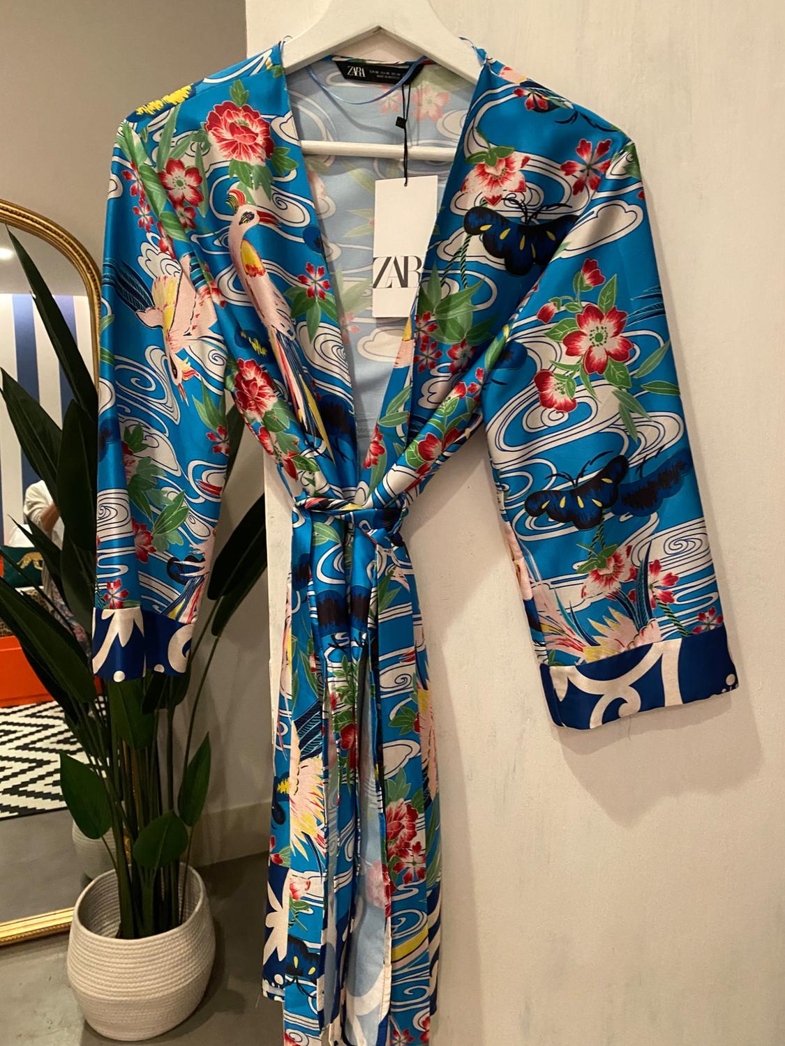 Blue dressing gown with a floral and bird print, hanging on a white hanger. Visible label with the text "ZARA". Background with a mirror and plant.
