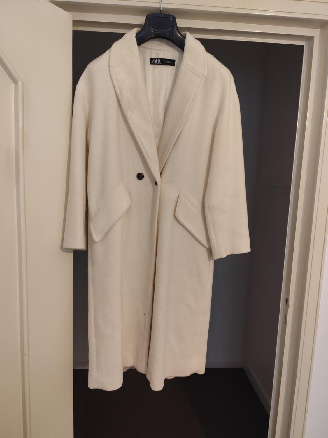 Long white coat hanging on a black hanger inside a door frame. Visible label with the text "ZARA".