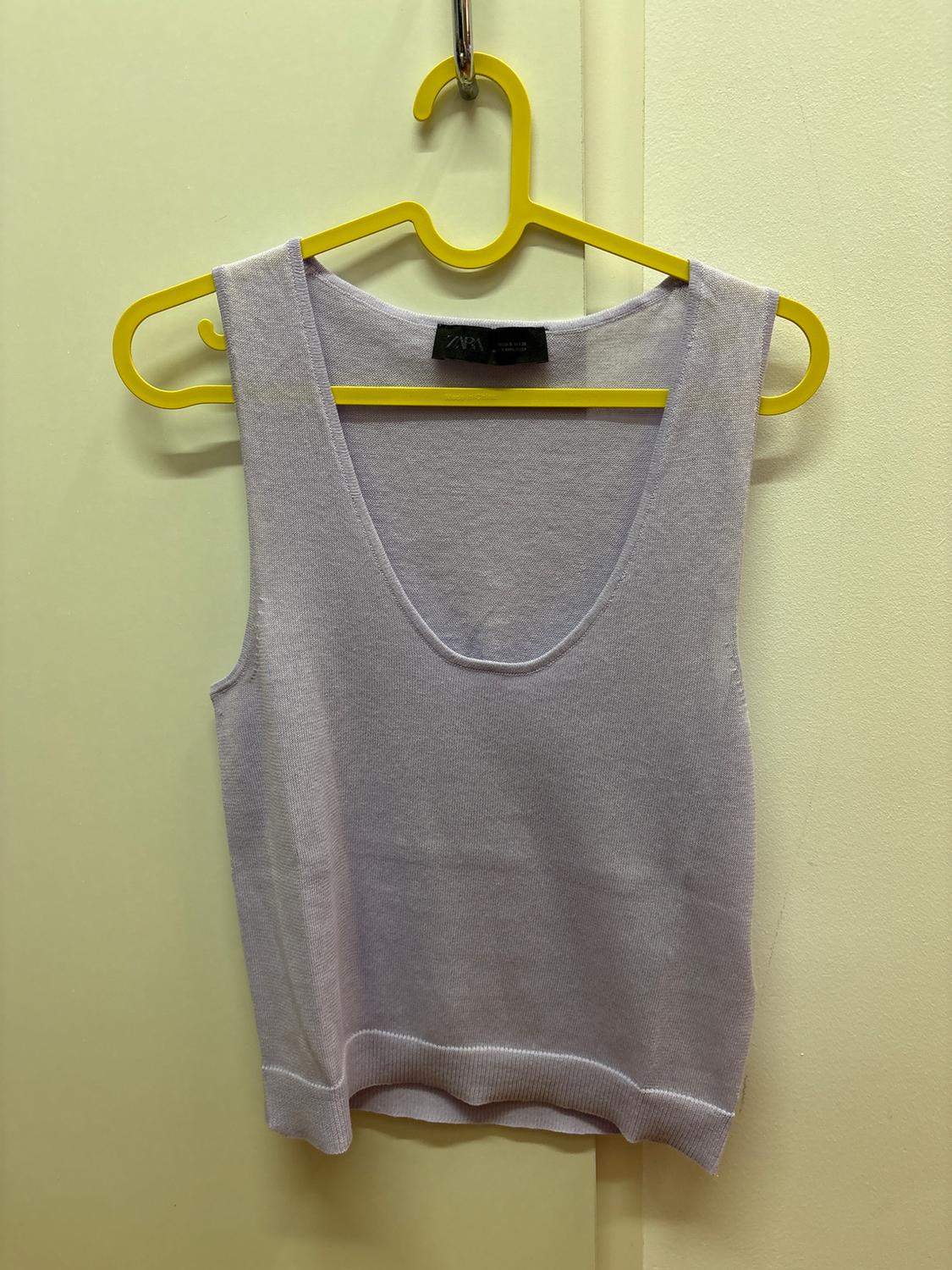 Light grey sleeveless top hanging on a yellow hanger inside a changing room. Label visible on the collar.