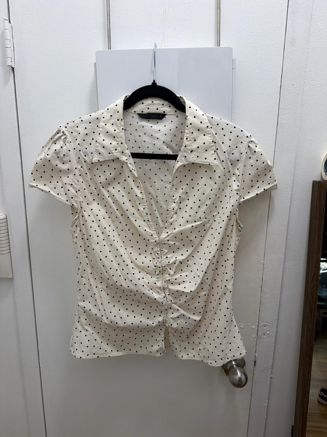 White blouse with black polka dots, short sleeves, a V-neck, and a gathered front with buttons, hanging on a hanger over a white door.