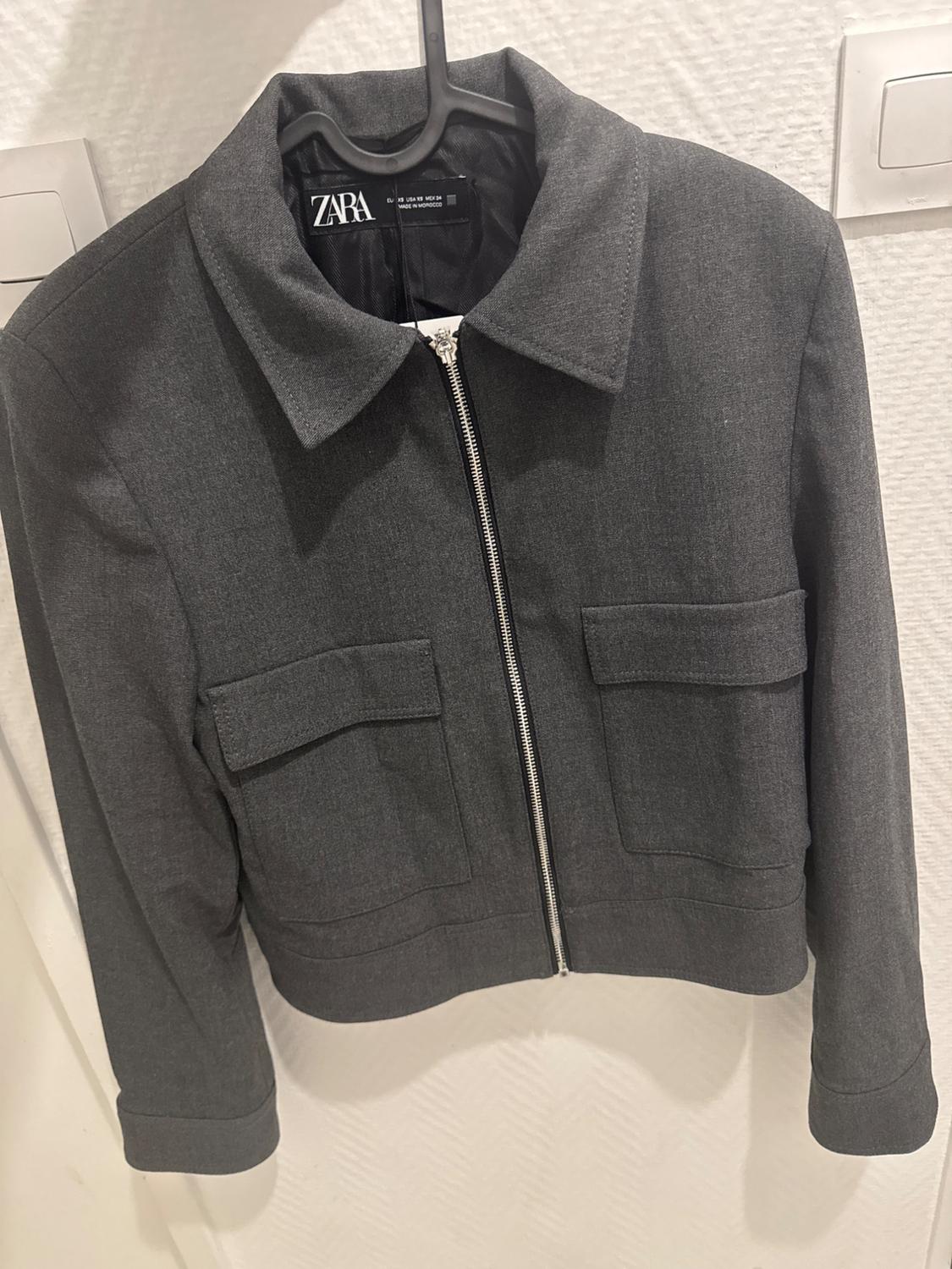 Grey jacket with a zip and front pockets, hanging on a black hanger. Visible label: "ZARA". Textured white background.