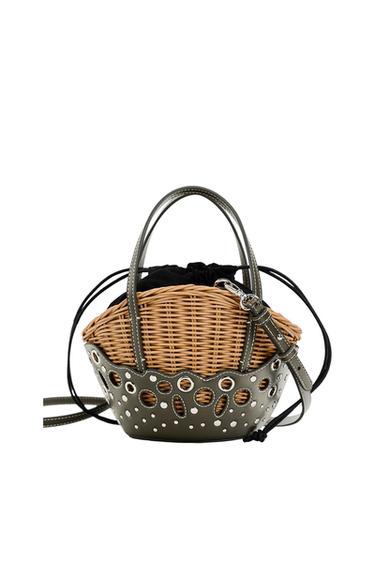 Wicker basket bag with an olive green base and handles. It features cutouts with metal appliqués and a black interior pouch with a drawstring closure.