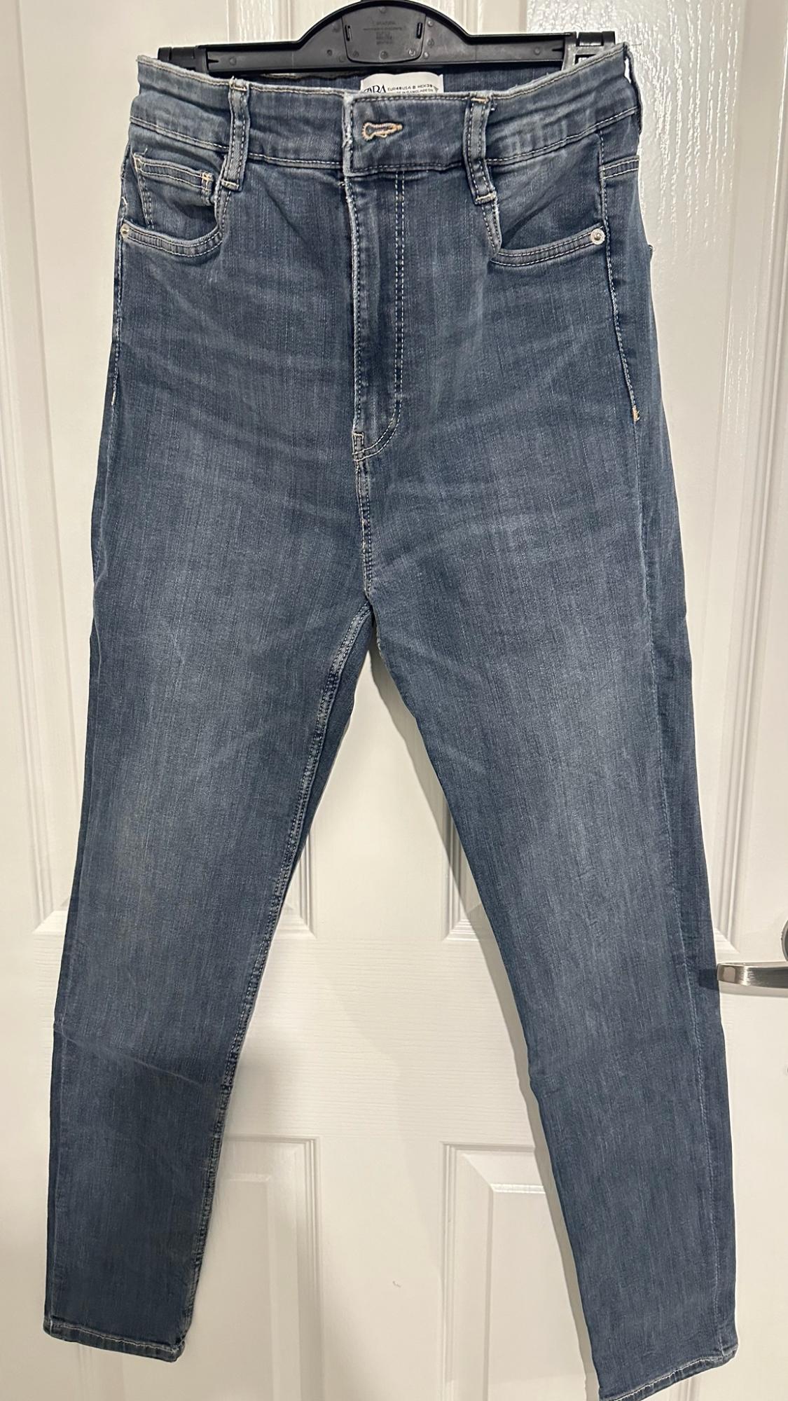 High-waisted blue jeans hanging on a black hanger in front of a white door.