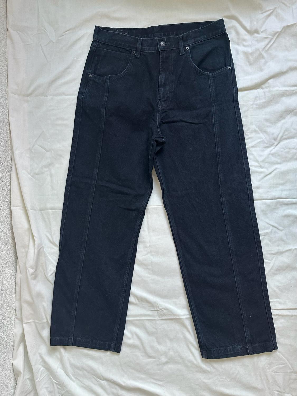 Black straight-leg jeans with front seams, laid out on crinkled white fabric in a casual style.