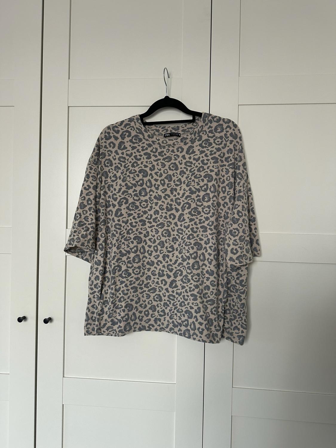 Γκρι oversized μπλουζάκι με animal print και κοντά μανίκια, κρεμασμένο σε μαύρη κρεμάστρα μπροστά από λευκές πόρτες ντουλάπας.