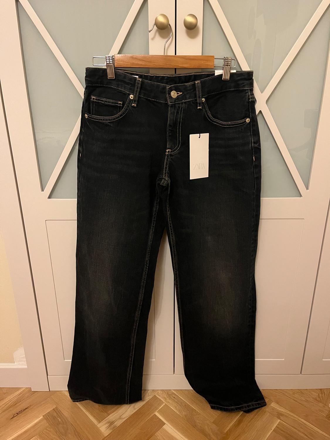 Dark jeans hanging on a wooden hanger in front of a white door. Label with the text ZARA. Herringbone wooden floor.