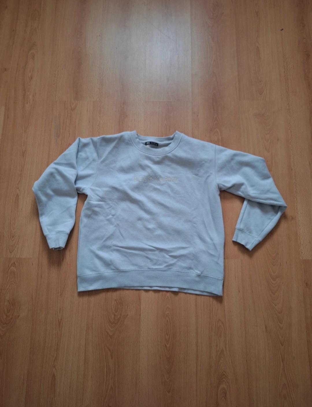 White sweatshirt with "DEAR KARMA" text on the chest, laid out on a light wooden floor.