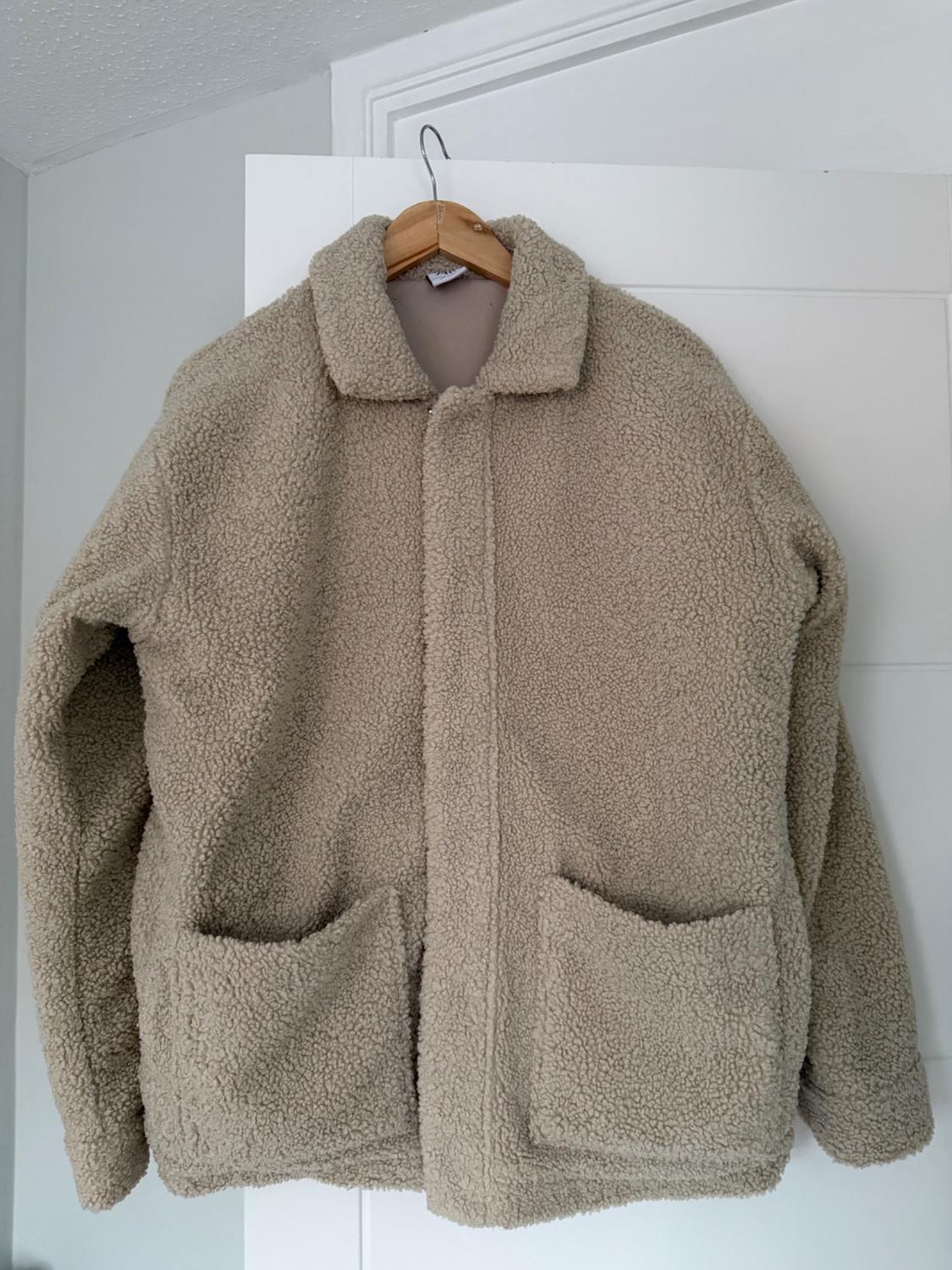 Beige jacket with a faux-fur effect on a wooden hanger over a white door. Design with a collar, hidden fastening and two large pockets.