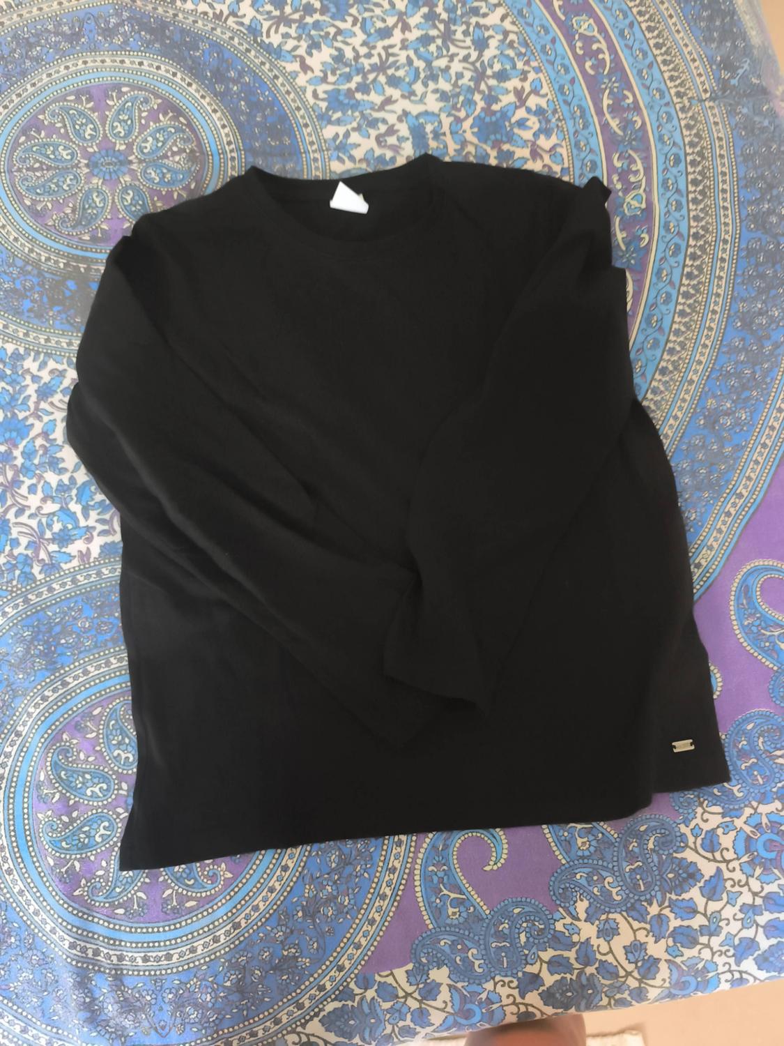 Basic black long-sleeved T-shirt laid out on fabric with a blue and purple pattern. It has a small metal badge at the hem.