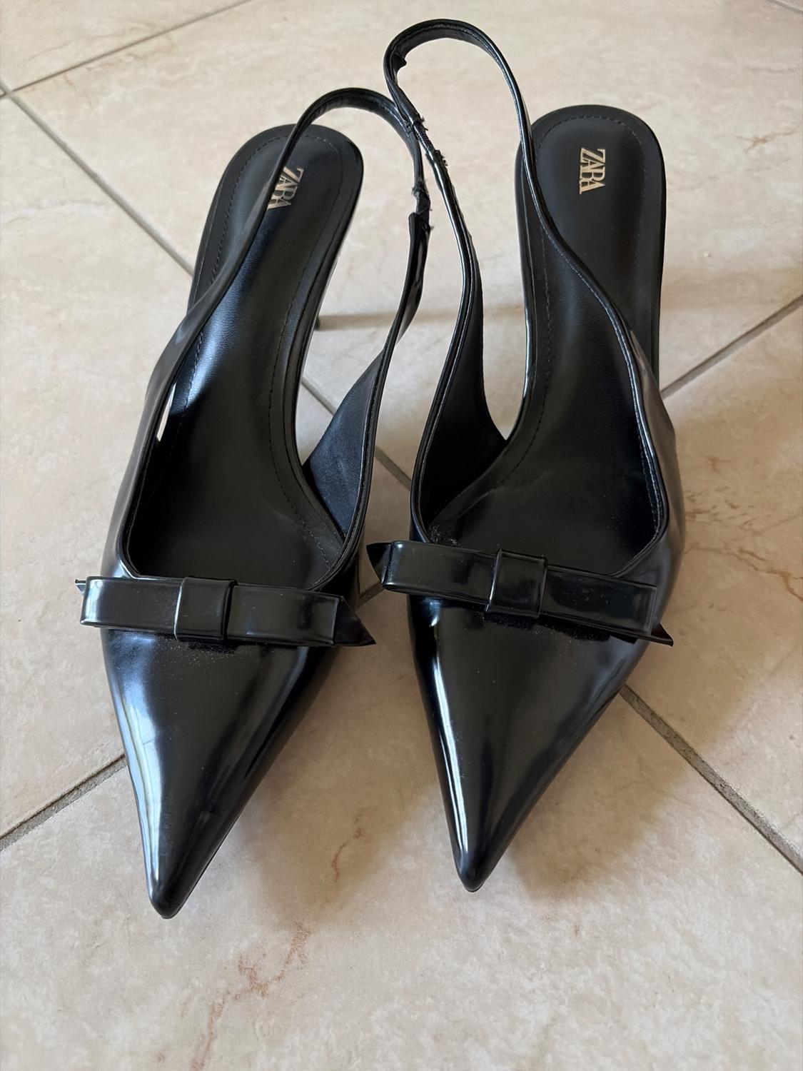 Black pointed-toe slingback shoes with bow detail on a tiled floor. ZARA text visible on the insoles.