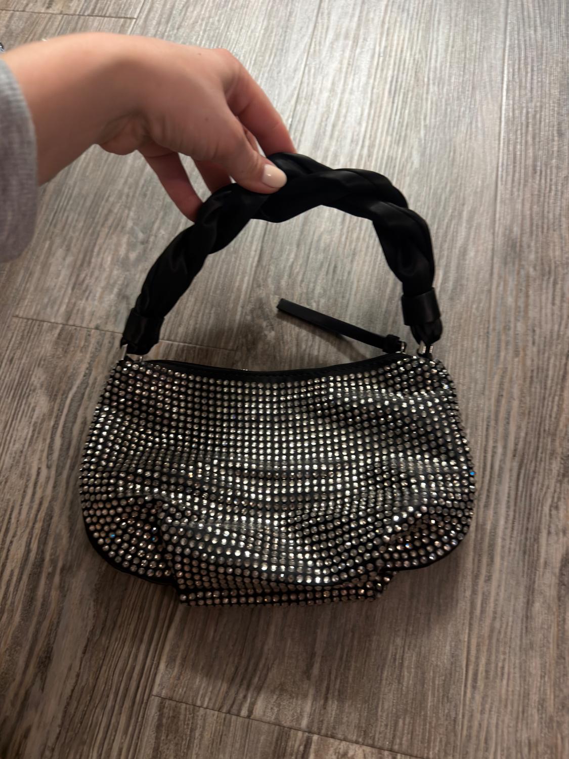 A small handbag adorned with sparkling rhinestones and a black braided handle, held by a hand over a light wooden floor.