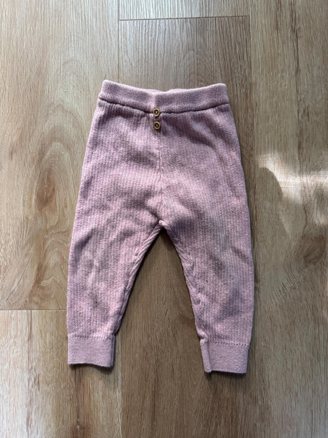 Light pink baby trousers with a ribbed texture and two buttons on the waistband, on a light wooden floor.
