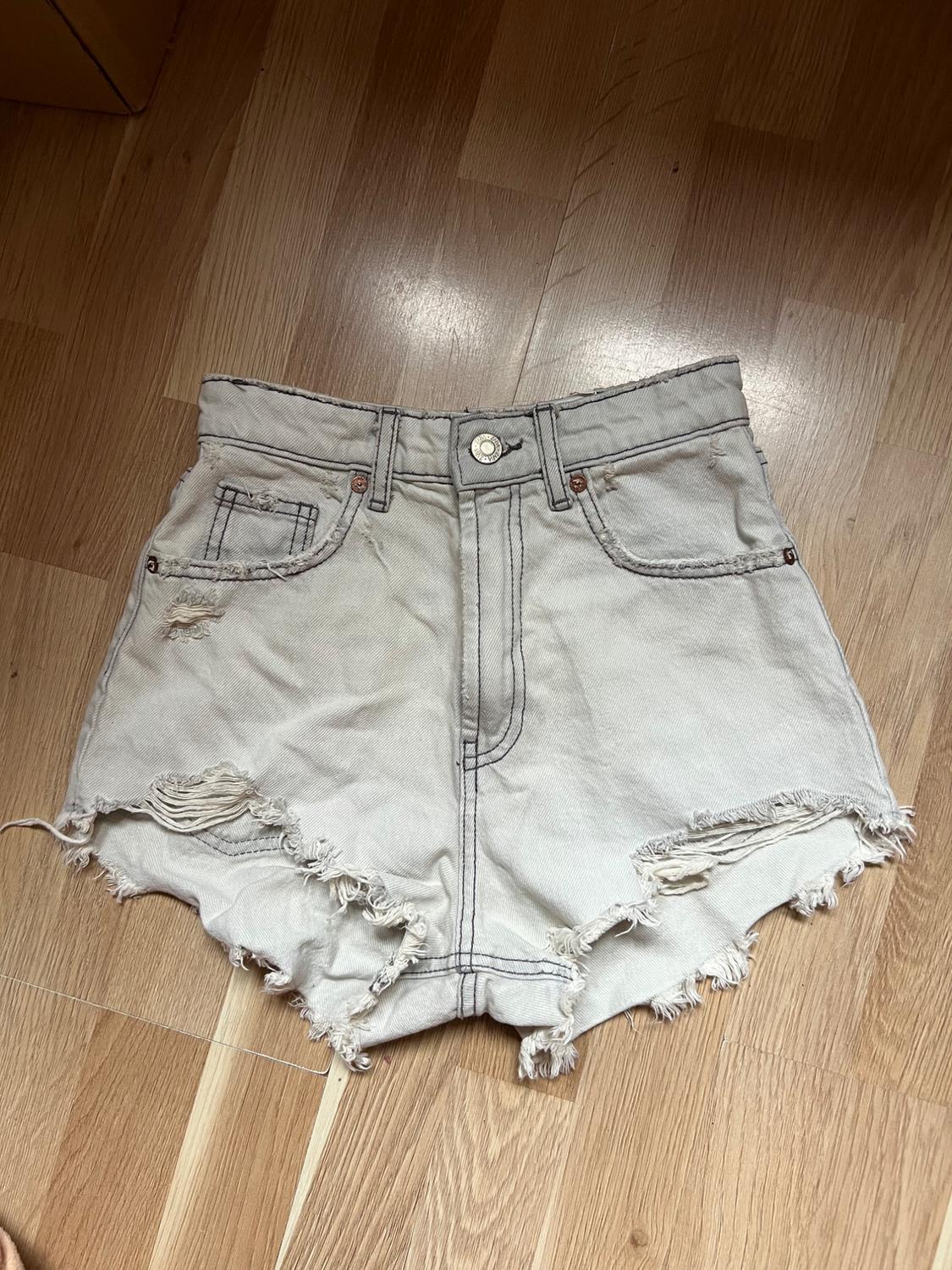 Light beige distressed denim shorts with frayed edges, front pockets, and a metal button, on a wooden floor.