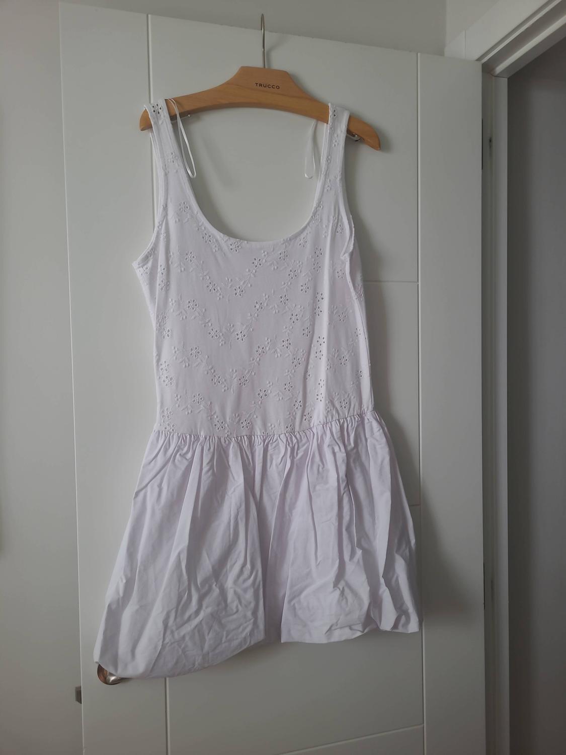 Sleeveless white dress with floral embroidery and a gathered skirt, hanging on a white door on a wooden hanger with the text TRUCCO.