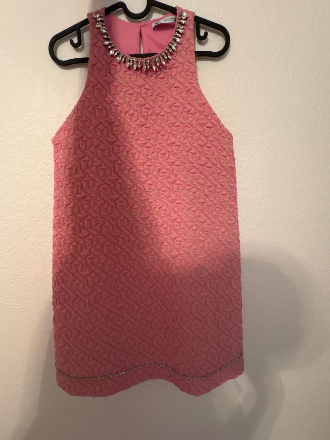 Sleeveless pink dress with geometric texture and jewel-embellished neckline, hanging on a black hanger.