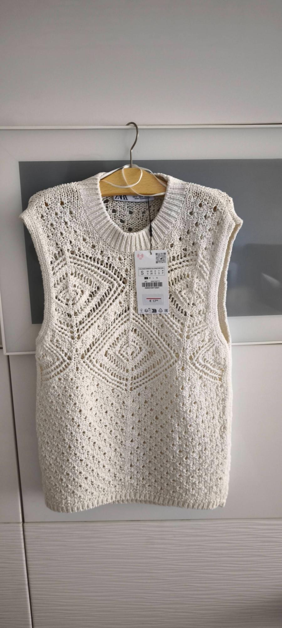 White knitted tank top with geometric design hanging on a wooden hanger. Visible tag with price of €17. Light grey background.