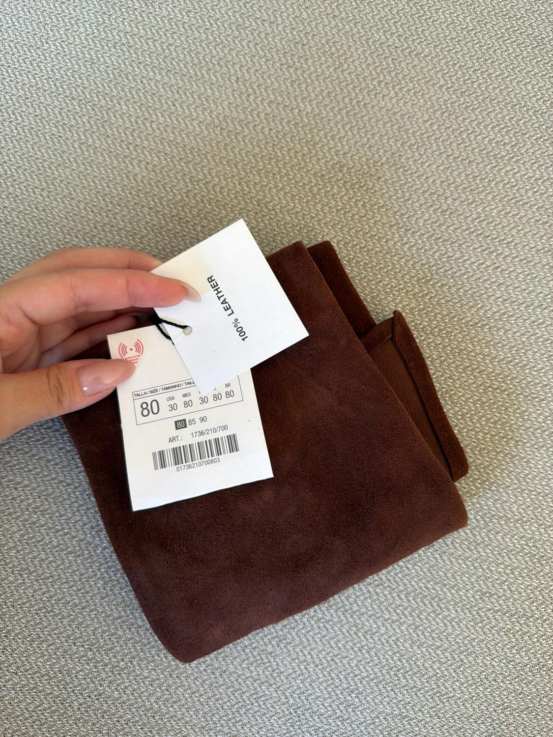 Hand holding a label on a brown garment. Text: 100% LEATHER, SIZE 80, USA 30, MEX 80, ART.: 1736/210/700, 01736210700803.
