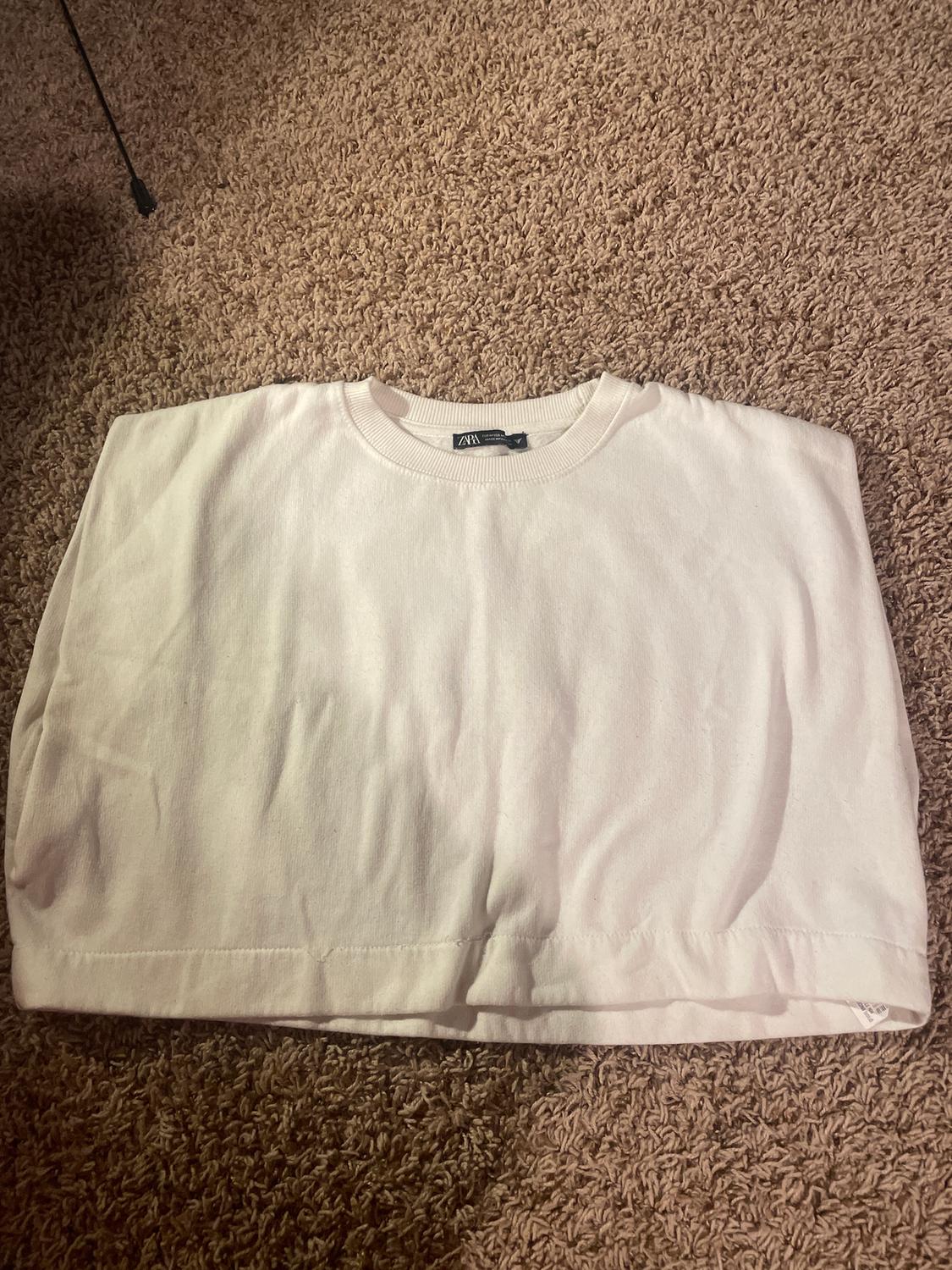 White, short-sleeved T-shirt folded on a brown rug. Visible label with text "ZARA".