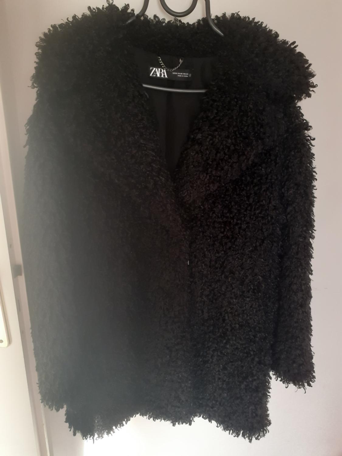 Black textured coat hanging on a hanger against a white wall. The inner label reads ZARA.