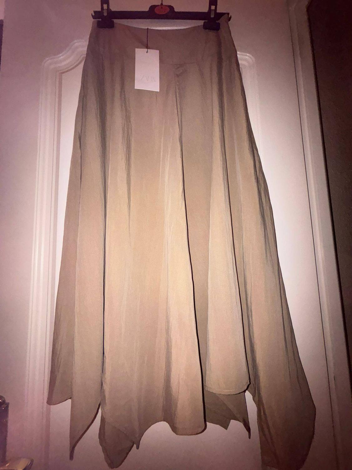 Beige long skirt with an asymmetric hem hanging on a black hanger over a white door. It has a label bearing the text ZARA.