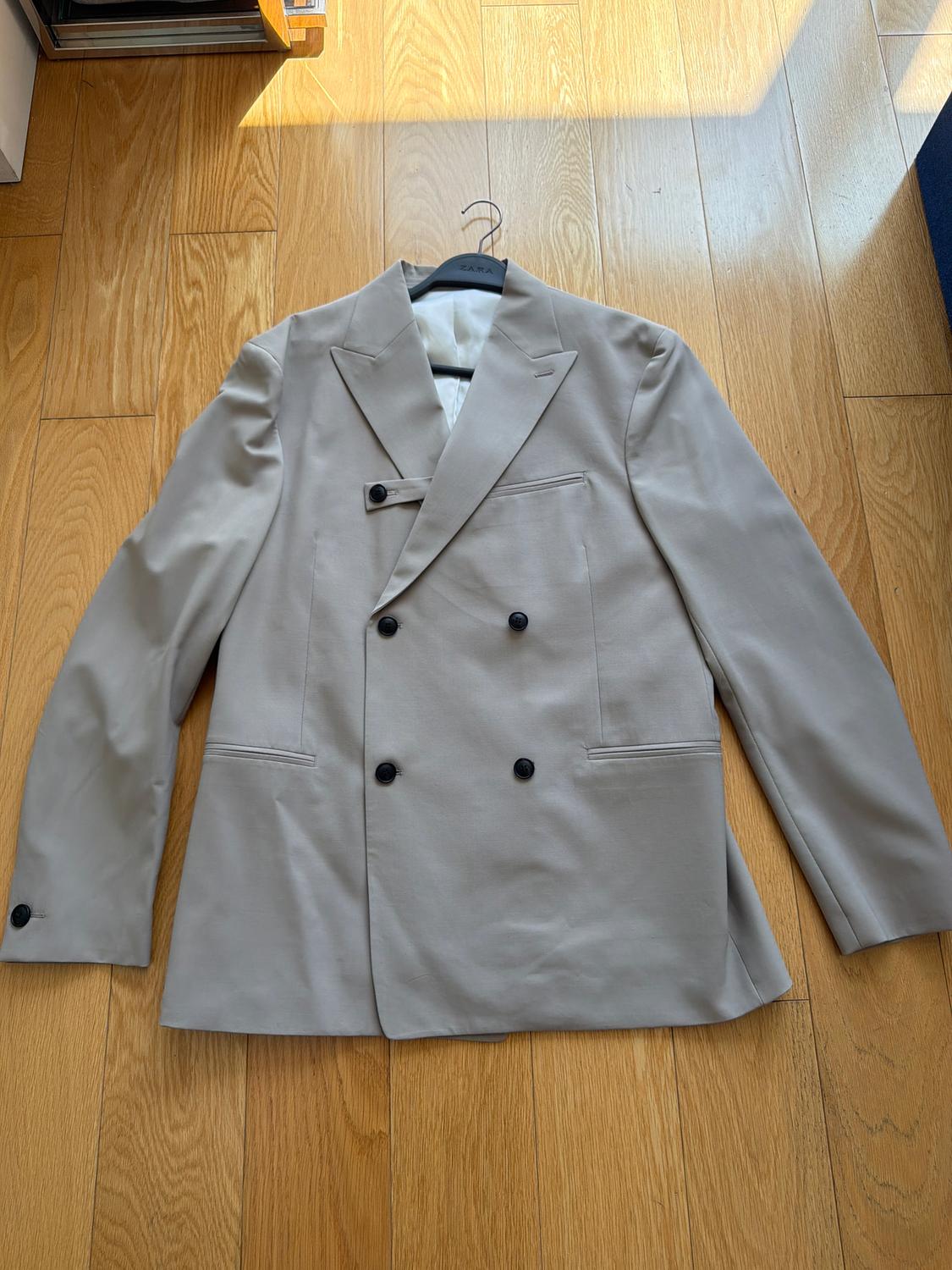 Beige double-breasted jacket with black buttons on a light wooden floor. Hanger with visible logo.