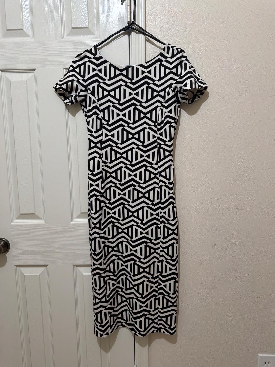 Short-sleeved dress with a geometric black-and-white pattern, hanging on a coat hanger on a white door next to a wall.