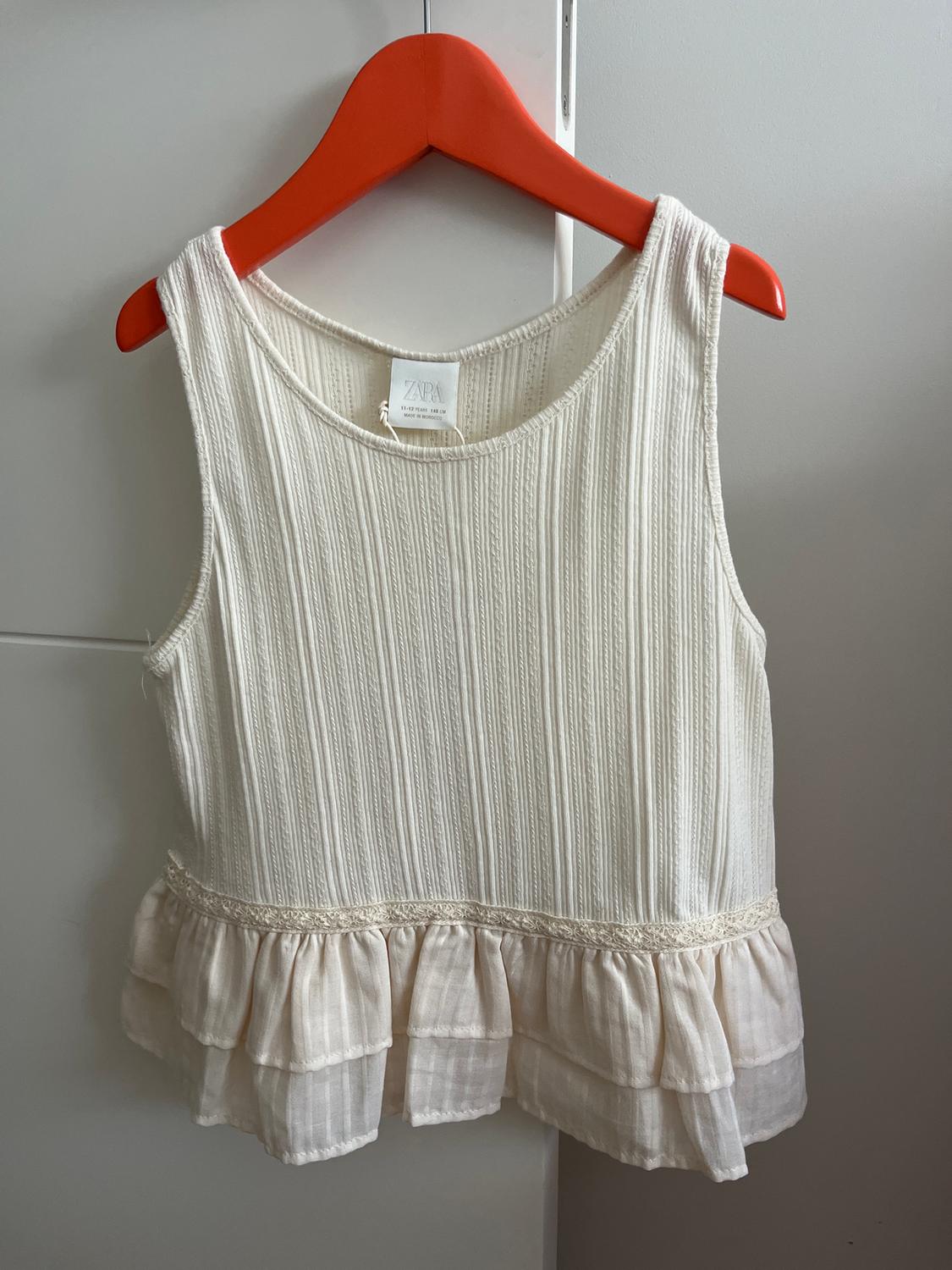 White sleeveless blouse with ruffle hem, hanging on a red hanger. Visible tag with text "ZARA".