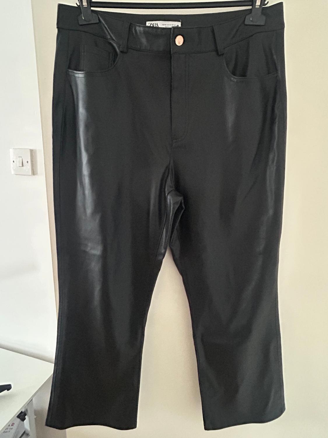 Black trousers hanging on a hanger, with a glossy finish and gold button. White background and visible plug socket on the left.