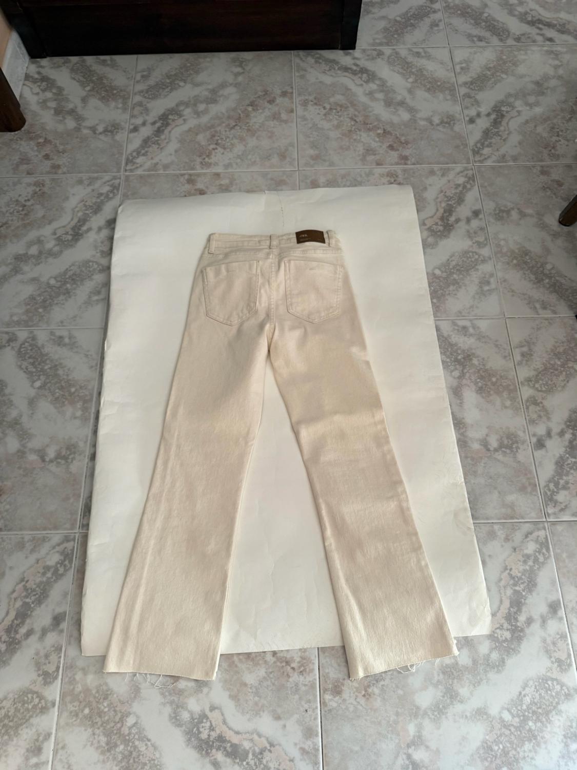 Beige denim jeans seen from behind with a frayed hem, laid out on white paper on a tiled floor. Relaxed atmosphere.