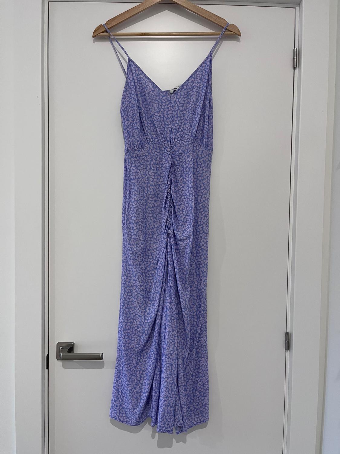 Lilac leaf-print dress with spaghetti straps and a gathered front, hanging on a wooden hanger on a white door.