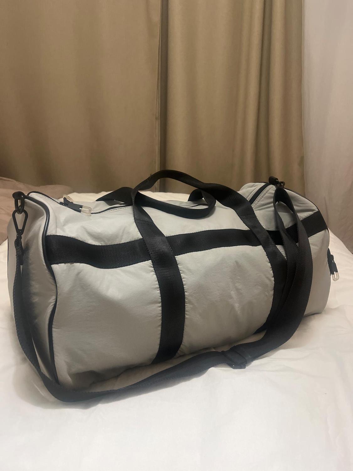 Light grey travel bag with black straps, resting on a white bed. Beige curtain background.
