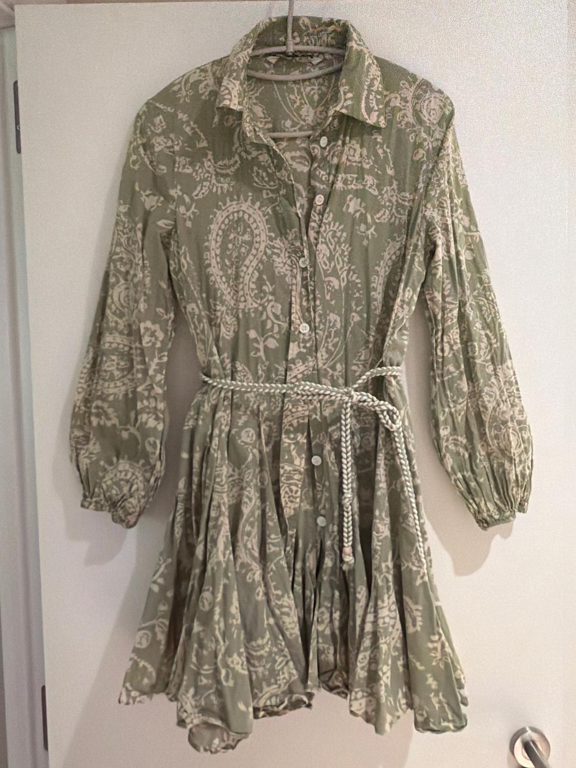 Green dress with a white floral print, long sleeves, and a braided belt, hanging on a hanger over a white door.