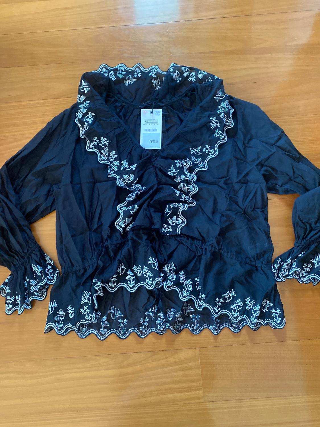 Black blouse with white embroidery on the collar, sleeves, and hem. Visible tag with a price of 79.90. Light wooden background.