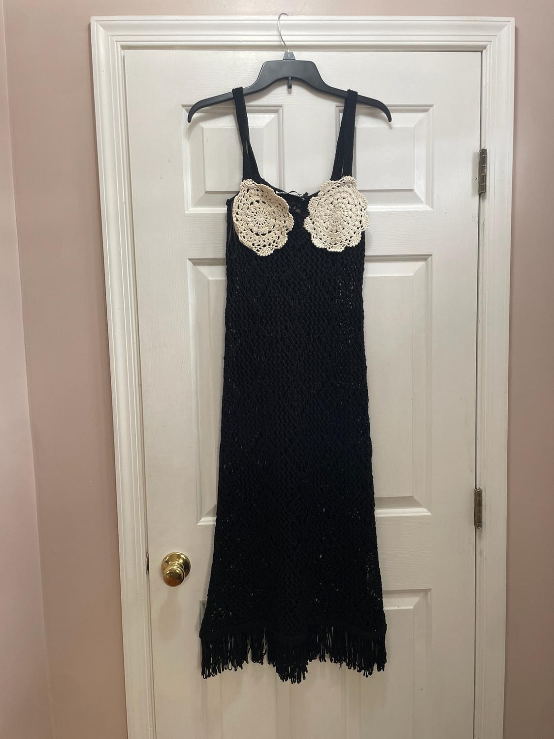 Black open-knit dress with white appliqués on the chest and fringe, hanging on a hanger over a white interior door.