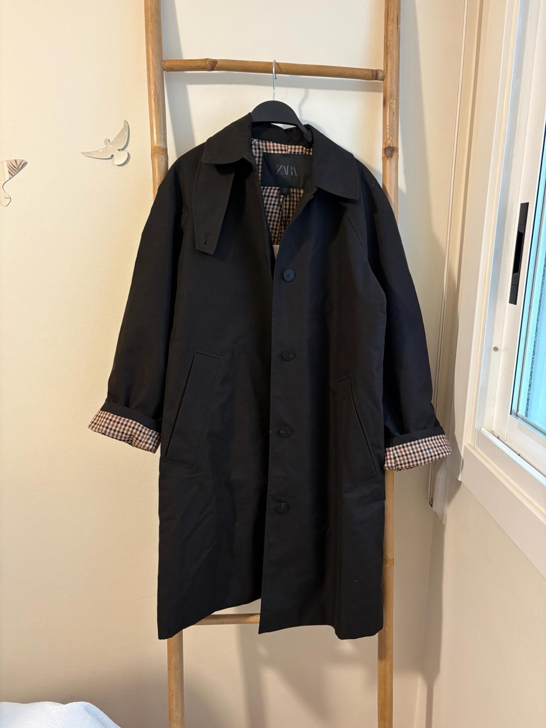 Black coat hanging on a wooden staircase. Shows tartan lining on the cuffs and collar, and the Zara label.