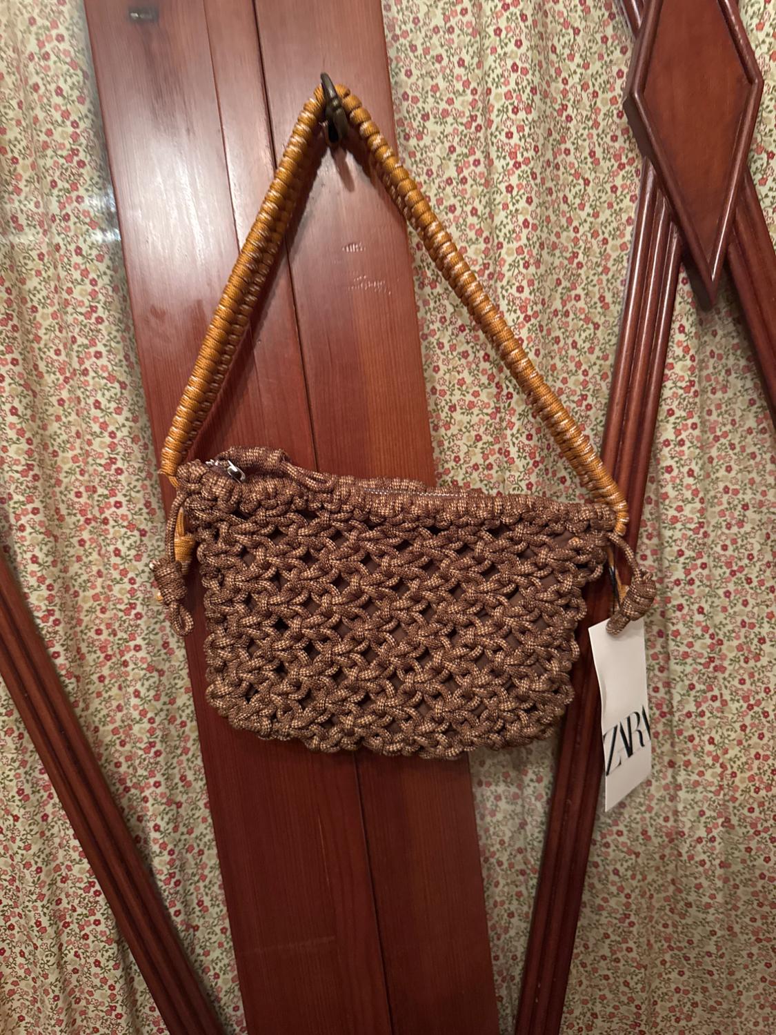 Brown knitted bag with wooden handle hanging on a wooden door. Fabric background with floral print. Visible label: ZVR.