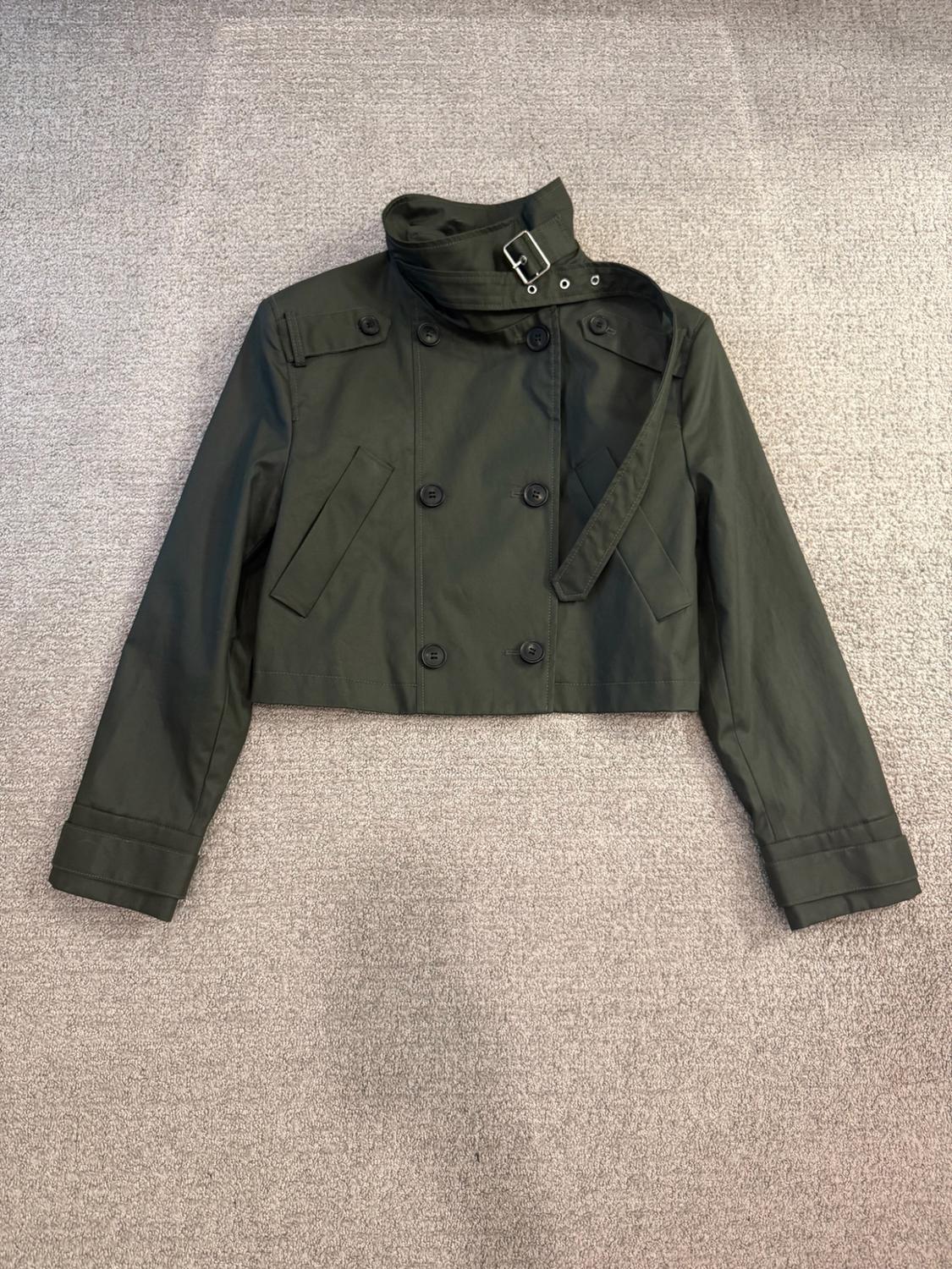 Short olive-green jacket with a double-breasted front, high collar with a buckle, and side pockets, arranged on a gray rug.