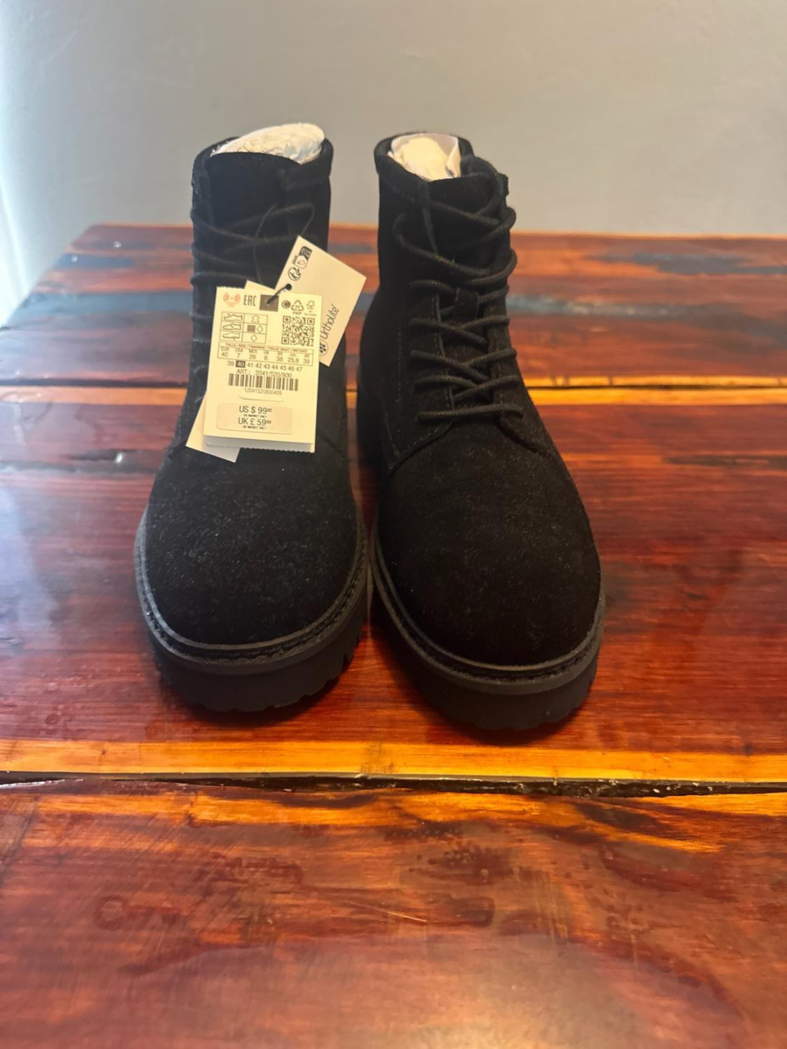 Black lace-up boots with thick soles on wood. Label with text: "Ortholite US $ 99.90 UK £ 59.99".