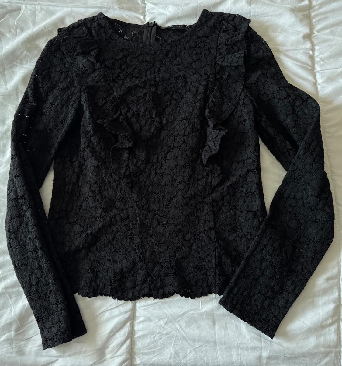 Black lace blouse with long sleeves and ruffles on the shoulders, on a wrinkled white surface.
