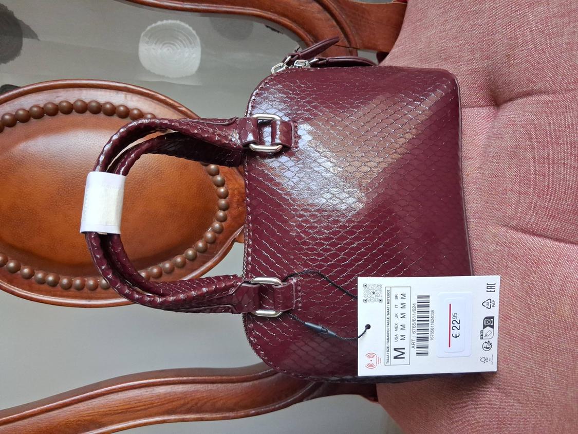 Τσάντα μπορντό γυαλιστερή με υφή φιδιού πάνω σε ξύλινη καρέκλα. Ετικέτα: M USA MEX UK IT BR M M M M M ART: 6765/611/624 € 22.95 EAC PAP.