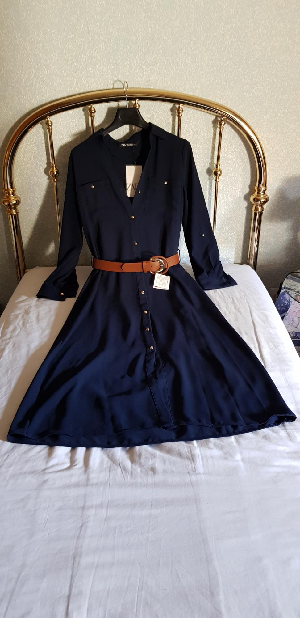 Navy blue dress with gold buttons and a brown belt on a bed with a gold headboard. Visible tag.