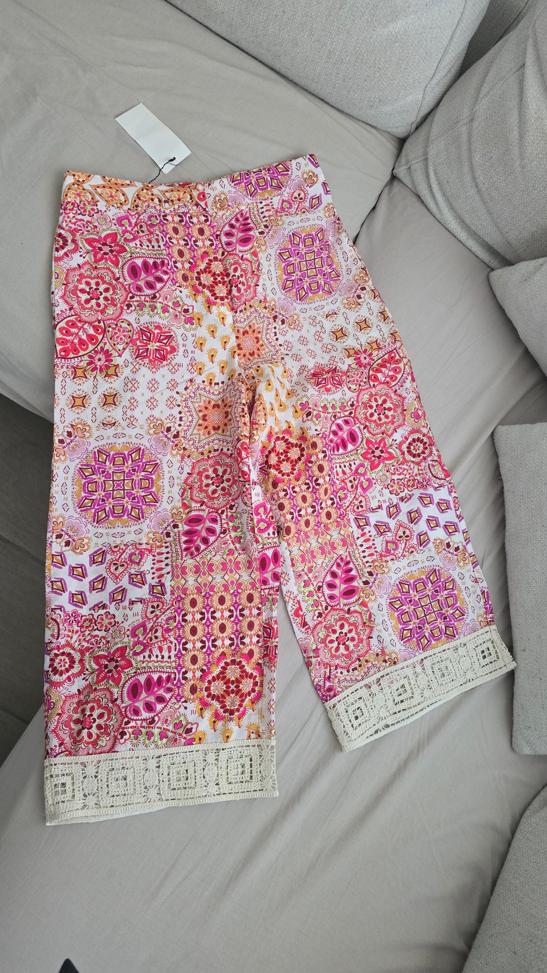 Printed trousers in pink and orange tones with floral details and white lace trim, on a light grey sofa.