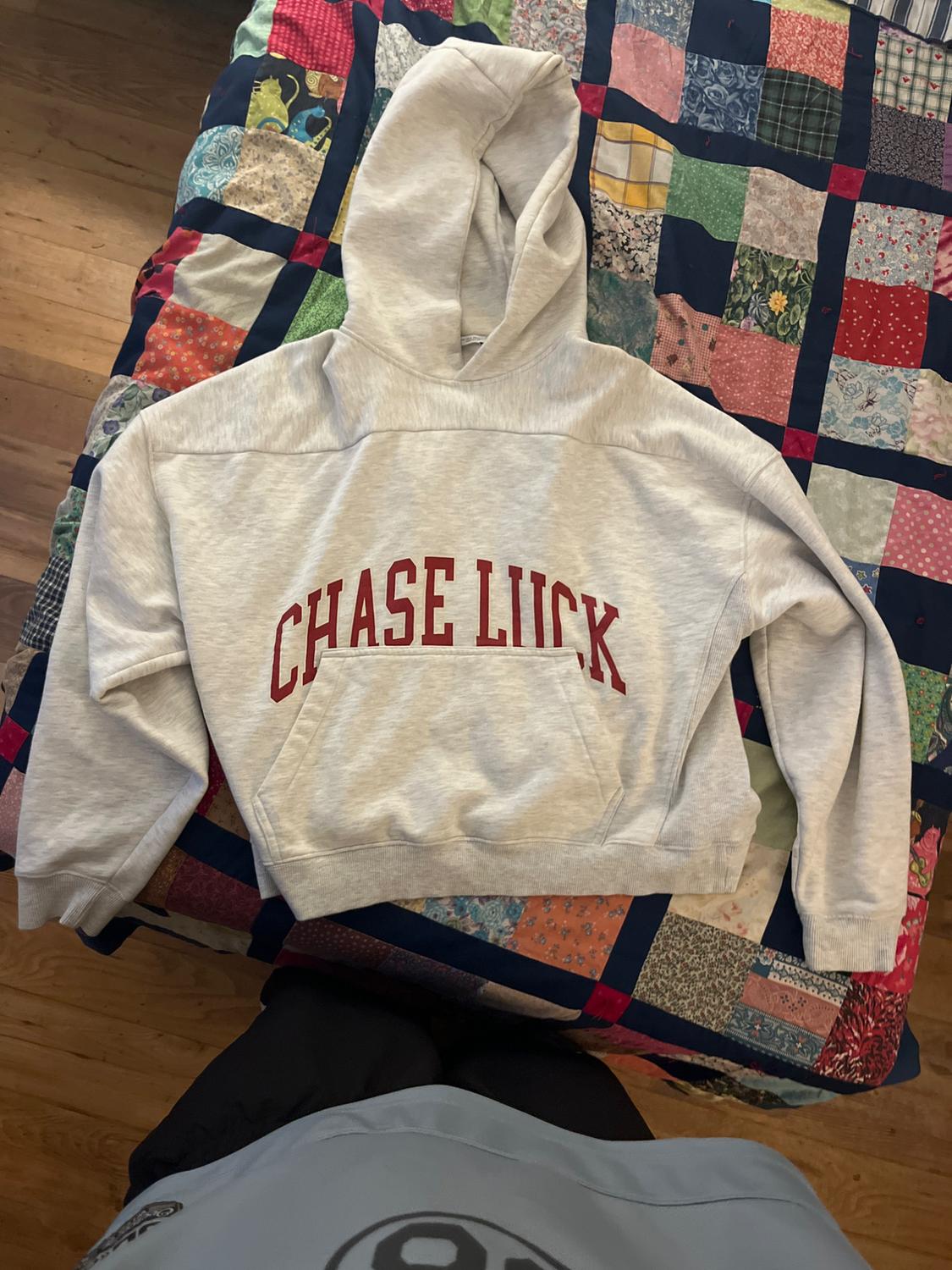 Grey hoodie with "CHASE LUCK" text in red, on a colourful patchwork quilt on a wooden floor.